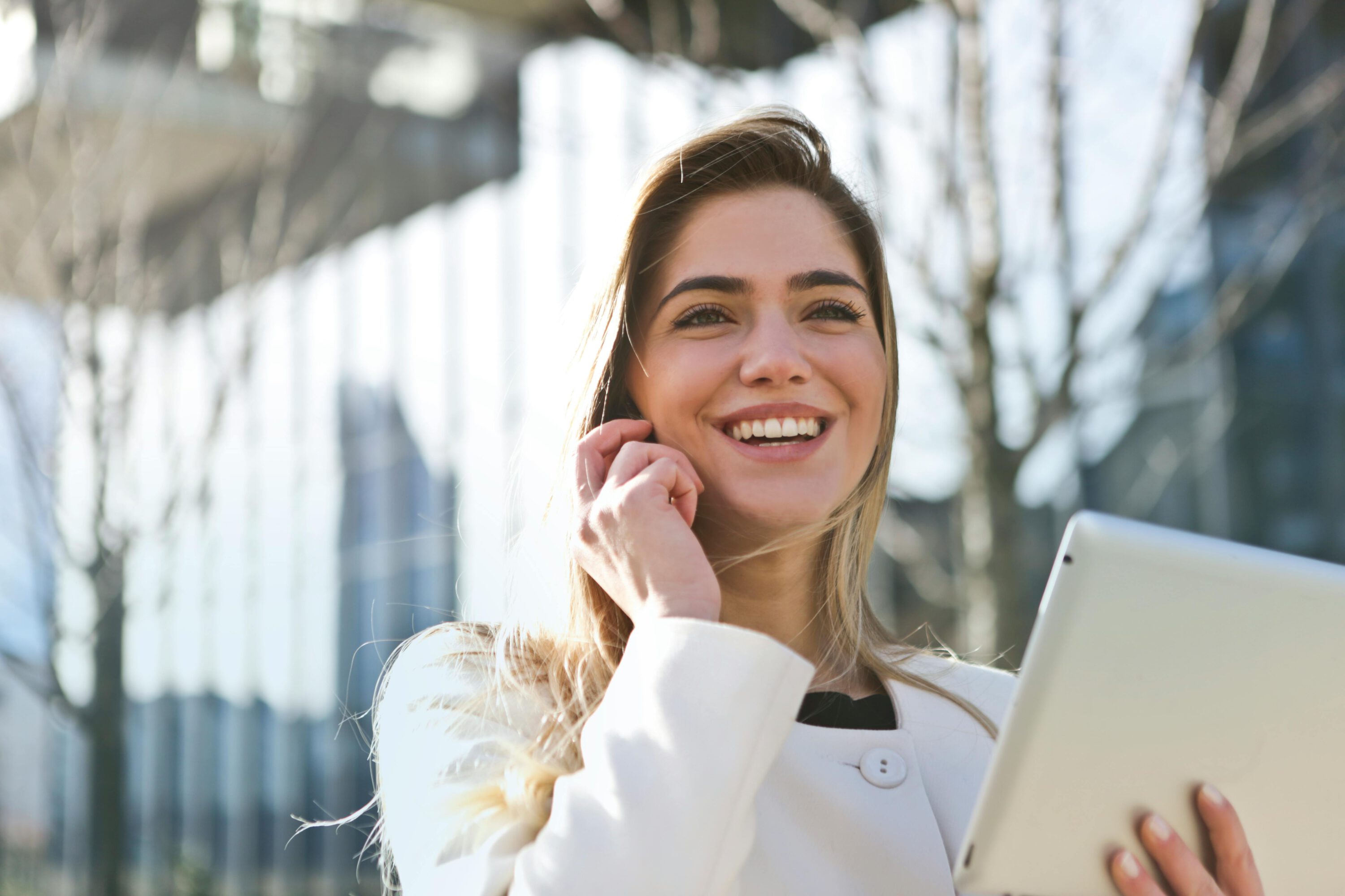 Une femme vêtue d'un blazer blanc, tenant une tablette dans ses mains, avec une expression concentrée, debout dans un environnement professionnel lumineux.